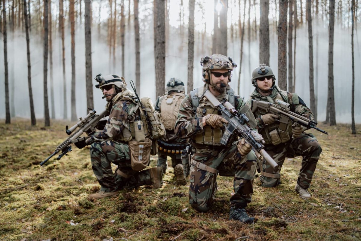Four fully equipped, middle-aged soldiers in camouflage uniforms form a line, ready to fire, aiming with their rifles. A military operation in action, a unit standing in a dense forest.