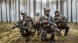 Four fully equipped, middle-aged soldiers in camouflage uniforms form a line, ready to fire, aiming with their rifles. A military operation in action, a unit standing in a dense forest.